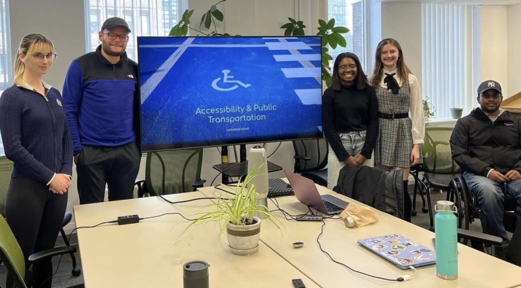 Four students, including Catherine, and alum, Igor, smiling next to the presentation screen at the Accessibility & Public Transportation U-TAAC discussion.